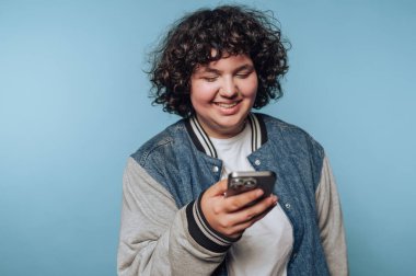 Kıvırcık saçlı, akıllı telefonlu bir genç, sıradan bir ortamda mavi bir arkaplan kullanıyor.