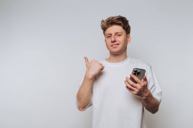 Genç adam akıllı telefonu tutarken gülümsüyor ve minimalist bir ortamda kamerayı işaret ediyor.