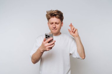 Genç adam telefonu umutlu bir ifadeyle kontrol ederken şans diliyor. Parlak, minimalist bir ortamda.