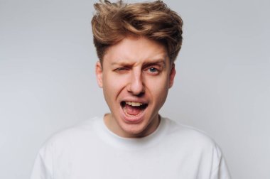 Heyecanlı genç adam, dağınık saçlı, neşeli ifade ederken, kapalı alanda komik bir yüz ifadesi takınıyor.
