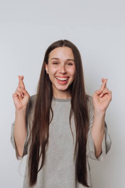 Umut dolu bir kadın büyük bir gülümseme ile parmaklarını çapraz yapar, beyaz arka planda beklenti ve heyecan ifade eder.