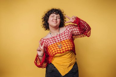 Renkli bir elbiseyle barış işareti gösteren genç bir kadın sarı arka planda neşe ve pozitifliği özetliyor.