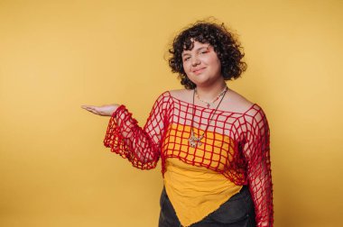 Kendine güvenen bir kadın, sarı arka planda cesur ve renkli bir elbiseyle hoş geldin jesti yapıyor.