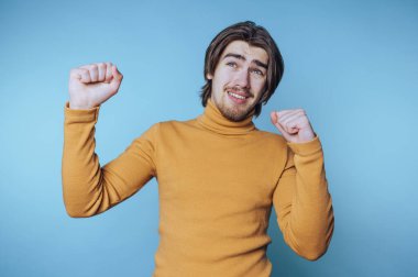 Canlı turuncu süveter giyen genç adam sevinç saçıyor, kolları mavi arka planda enerji dolu kutlama yapıyor.