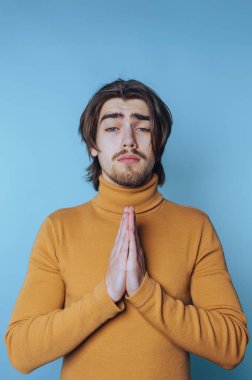 Balıkçı yakalı genç adam ellerini dua etmek için birleştirir. Mavi arka planda umut ve samimiyet gösterir.