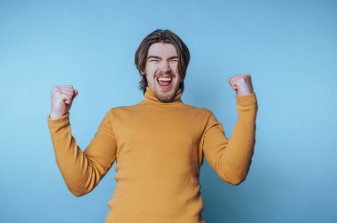 Boğazlı kazaklı genç adam sevinçle haykırıyor, başarısını mavi arka planda yumruklarını kaldırarak kutluyor.