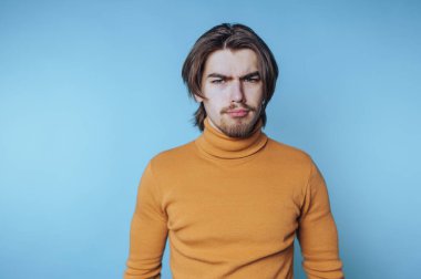 Boğazlı kazaklı genç adam çatık kaşlı, kararlılığını yakalıyor ve mavi arka plana odaklanıyor.