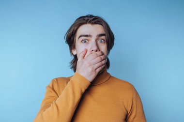 Genç adam, mavi arka planda poz verirken şaşkınlık ve şaşkınlık içinde ağzını kapatır.