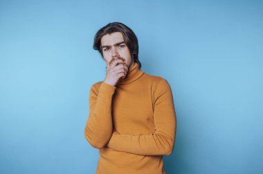 Genç adam düşünceli bir duruşta duruyor, elleri çenesinde, derin fikirleri ve mavi arka plandaki kavramları yansıtıyor.