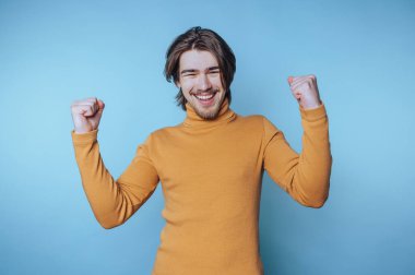 Genç adam, mavi arka planda pozitif enerji ve neşe saçan coşkulu bir gülümseme ve havaya kaldırılmış yumruklarla kutluyor.