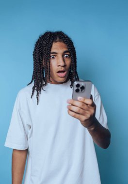 Rastalı genç adam, mavi arka planda duygularını sergileyen akıllı telefonuna bakarken şaşkınlığını dile getiriyor.