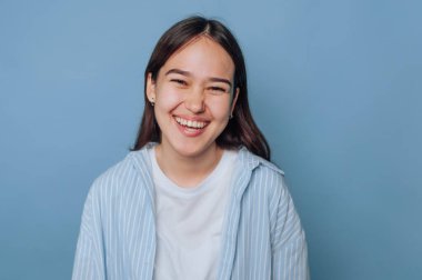 Gülümseyen genç kadın neşeli kişiliğini sergiliyor, açık mavi arka planda mutluluk saçıyor.