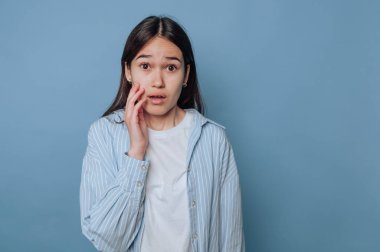 Genç bir kadın, eli yüzünde endişeli bir bakış sergiliyor, açık mavi arka planda endişe ya da şaşkınlık hissi taşıyor.