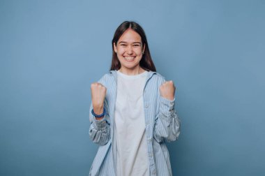 enerjik genç kadın yumruklarını kaldırarak sevinç ifade ediyor, kutlamaları yakalıyor ve açık mavi arka planda zafer kazanıyor.