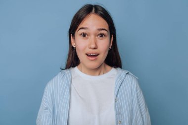 Genç bir kadın açık mavi arka planda kameraya bakarken şaşkınlık ifadesi sergiler.