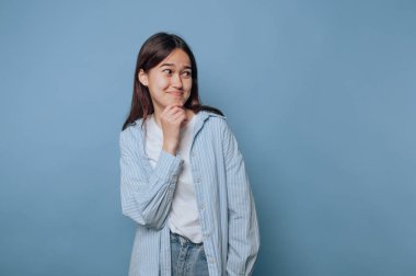 Genç bir kadın, açık mavi arka planda muzip bir sırrı somutlaştıran bir gülümseme ile çenesine neşeyle dokunur.