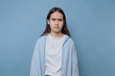 Genç bir kadın hüzünlü bir ifadeyle hoşnutsuzluk gösterir, açık mavi arka planda bir anlık yansıma ve hayal kırıklığı yakalar.