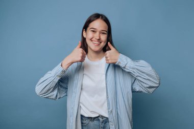 Mutlu genç kadın, olumlu bir jest paylaşıyor. Açık mavi arka planda pozitif bir hava ve heyecan yayıyor.