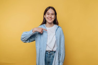 Genç bir kadın gülümsüyor ve parlak sarı arka planda kendine işaret ediyor. Kendine güvenini ve güvenini ifade ediyor.