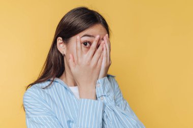 Kadın elleriyle yüzünü kapatıyor, anksiyetesini ve rahatsızlığını canlı sarı arka planda ifade ediyor.