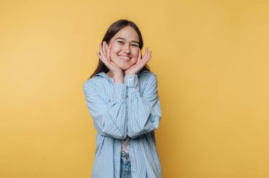 Gülümseyen genç bir kadın heyecanla yüzünü kavrıyor, canlı sarı bir arka plana kurulmuş, sıcaklık ve neşe hissi uyandırıyor.