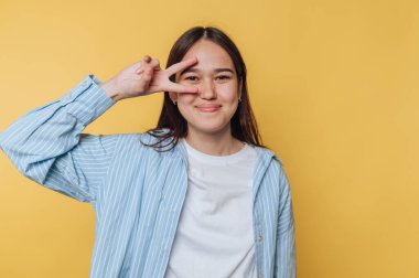 Neşeli genç bir kadın, hayat dolu sarı bir arka planda nazik bir gülümsemeyle barış işareti yapıyor.