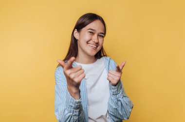 Mutlu genç bir kadın, canlı sarı arka planda gülümserken iki parmağıyla neşeli bir şekilde işaret ediyor.