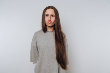 Düşünceli genç bir kadın hafifçe somurtuyor, beyaz arka planda kameraya bakarken kafası karışık bir yüz ifadesi sergiliyor.