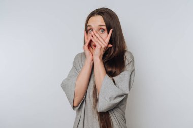 Korkulu kadın elleriyle yüzünü gizler, beyaz arka planda derin bir şok ve endişe taşır.