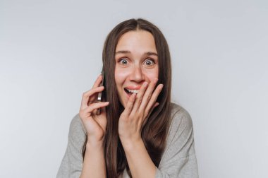 Kadın beyaz arka planda yüzüne yakın bir akıllı telefonu tutarken neşeyle ışıldıyor.