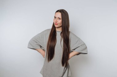 Kendine güveni tam genç bir kadın, elleri kalçasında duruyor, beyaz arka planda kendine güvenen ve şık bir tutum sergiliyor.