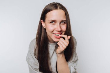 Genç bir kadın şakacı bir şekilde parmağını ısırıyor, beyaz arka planda bir entrika hissi uyandıran cilveli bir gülümseme ekliyor.