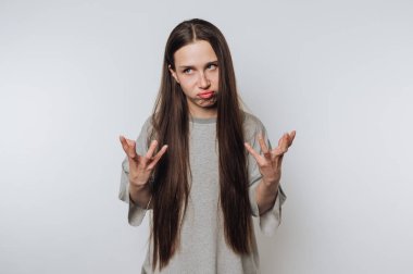 Hüsrana uğramış ifadesi ve kaldırılmış elleri olan genç bir kadın beyaz arka planda kafa karışıklığını resmediyor