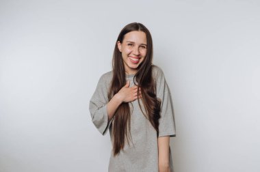 Genç bir kadın elini kalbine koyar gülümseyerek, minnettarlığı ve sıcaklığı simgeler beyaz arka planda.