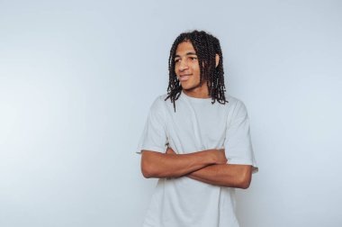 Kendine güvenen genç adam kollarını kavuşturmuş, düz beyaz bir tişört giyiyor ve beyaz arka planda düşünceli bir ifade sergiliyor.