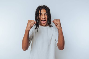 Beyaz tişörtlü genç adam heyecan dolu ışıldıyor, yumruklarını beyaz arka planda zaferle kaldırıyor.