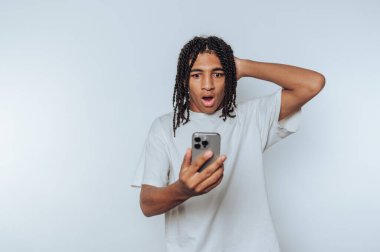 Genç adam, beyaz arka planda şok olmuş bir ifade göstererek akıllı telefonuna bakarken şaşkınlıkla tepki veriyor.