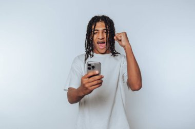 Genç adam akıllı telefonuna bakarken heyecan gösteriyor. Beyaz arka planda zaferin ya da başarının sevinç dolu bir anını gösteriyor.