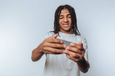 Genç adam akıllı telefonuyla meşgulken büyük bir gülümseme sergiliyor, beyaz arka planda bir anlık keyfi canlandırıyor.