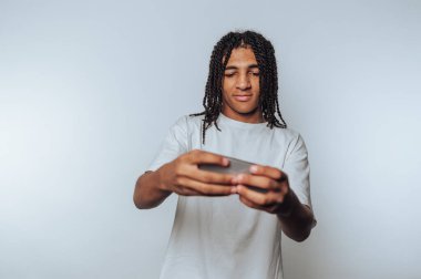 Genç, mobil cihazına odaklanıyor, oyuna odaklanıyor, konsantrasyon ve boş zamanlarını beyaz arka planda geçiriyor.