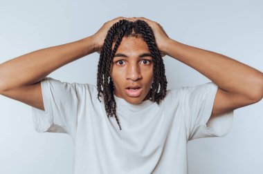 Şaşırmış genç adam inançsızlıkla başını tutuyor, güçlü duyguları yüz ifadeleri ve beyaz arka planda vücut dili ile aktarıyor.