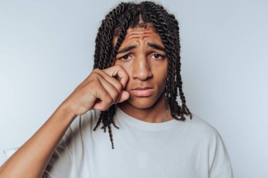 Kederli genç adam, beyaz arka planda kişisel yansımanın ve kırılganlığın özünü yakalayan nazik bir jestle gözlerini siliyor.