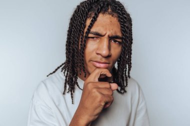 Genç adam düşünceli bir ifade ve jest sergiliyor, sanki derin bir düşüncelere dalmış, beyaz arka planda merak ve tefekkür sergiliyor.