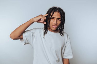 Saçları örgülü genç adam beyaz arka planda şakağına işaret ederken düşünceli bir ifade sergiliyor.