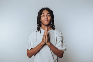 Genç adam dua ederken ellerini birbirine kenetliyor, beyaz arka plandaki umut ve yansımayı gösteren sakin bir ifade sergiliyor.