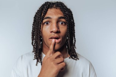 Genç adam, beyaz arka planda bir merak ve merak duygusu tasvir ederek, dudaklarında bir parmak ile şaşkın görünüyor