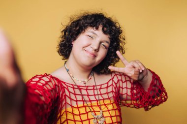 Neşeli genç kadın barış işareti ile poz veriyor, yaratıcı stilini ve sarı arka planda canlı kişiliğini sergiliyor.