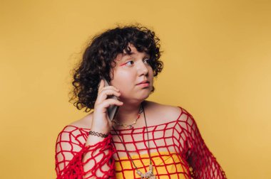 Genç adam düşünceli bir şekilde yana bakıyor, sarı arka planda tuhaf bir jest yaparken elinde bir telefon tutuyor.