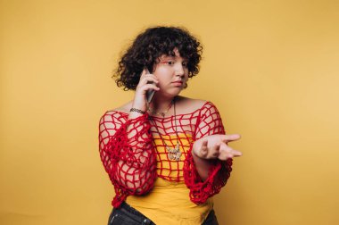 Genç bir kadın telefonuyla konuşurken şaşkın görünüyor. Sarı arka plandaki karışıklığı yansıtıyor.