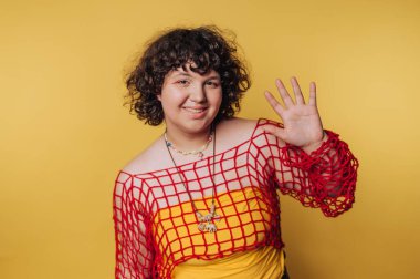 Genç kadın gülümserken el sallıyor, sarı arka planda hoş ve neşeli bir atmosfer olduğunu gösteriyor.